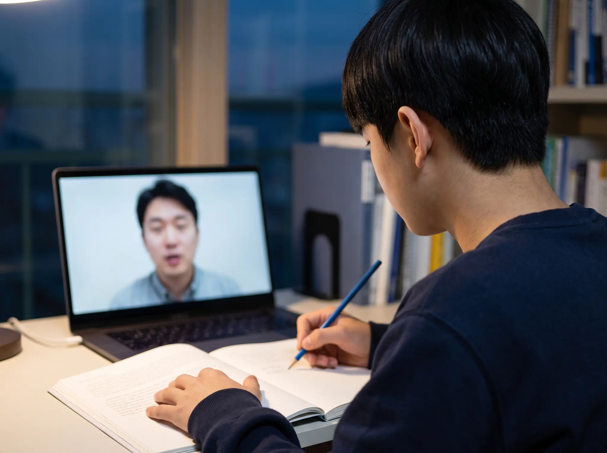 저녁 공부방에서 고등학생이 노트북 화면 속 선생님과 1:1 화상 수업을 하며 펼친 책에 파란 연필로 적는 어깨 너머 모습 — 고3 수학 과외 시범 수업